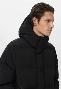 Chaqueta negra acolchada con un alto cuello y una gran capucha, con una textura suave y un diseño minimalista. Detalle de cierre frontal con botones.