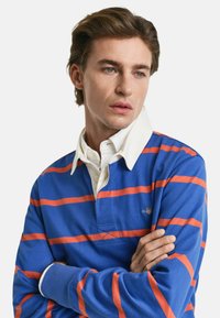 Blå og oransje stripet polo skjorte med hvit krage, med en knappestolpe og en logo på venstre bryst. Glatt tekstur.