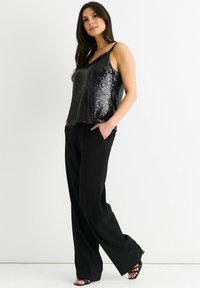 Black sequin camisole top with adjustable straps paired with loose-fitting black trousers. Model stands with hands in pockets, wearing strappy sandals.