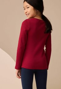 Haut rouge à manches longues avec une texture côtelée, présentant une large encolure et des manches ajustées, accompagné de leggings navy. Fond neutre.