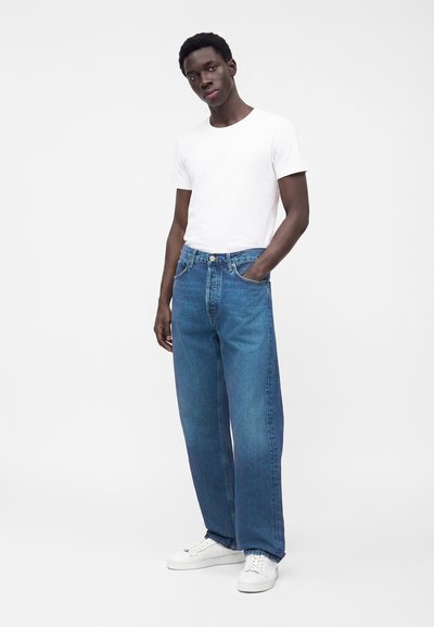 Homme portant un t-shirt blanc, un jean bleu à jambes larges et des baskets blanches, debout avec une main dans la poche sur un fond uni.
