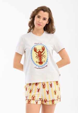 Mujer con camiseta blanca con una gran langosta roja y texto, combinada con pantalones cortos amarillos a rayas con varias langostas rojas.