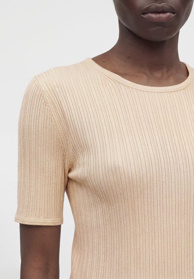 Κοντινό πλάνο ενός ατόμου που φοράει ένα μπεζ ριπ κοντομάνικο μπλουζάκι με στρογγυλή λαιμόκοψη σε μονόχρωμο φόντο.