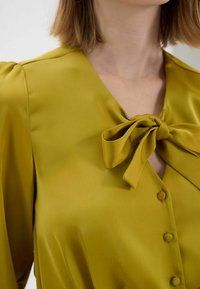 Blouse en satin vert olive avec un nœud au niveau du col, des manches bouffantes et des détails de boutons sur le devant. Texture lisse.