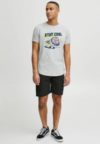 Szary bawełniany t-shirt z niebieskim i żółtym graficznym napisem "STAY COOL" oraz ilustracją; w połączeniu z czarnymi szortami i czarnymi sneakersami.
