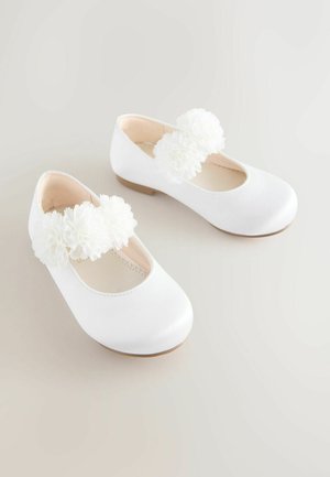 Baltie satīna baleta kurpes ar divām lielām baltām auduma ziedu rotājumiem katrā siksniņā, noapaļota purngala un zema, gaiši brūna zole.