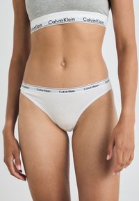 Biele bavlnené tangá s čiernym pásom s logom Calvin Klein. Mäkka textura, minimálny dizajn. Viditeľný je aj šedý crop top so podobným brandingom.