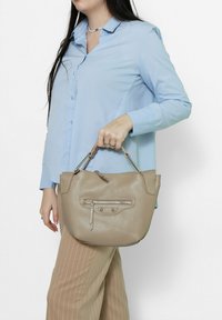 Beige leren handtas met een gestructureerde afwerking, bovenhandvatten en een voorvak met rits. Model houdt de tas vast terwijl hij een lichtblauw shirt draagt.