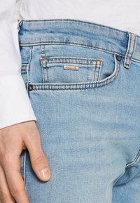 Jean en denim bleu clair avec un aspect usé, présentant des coutures marron et une étiquette "BOSS" sur la poche arrière. Main reposant sur la cuisse.