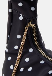 Czarna torba nylonowa w białe kropki, z złotym zamkiem błyskawicznym i detalem łańcucha. Widoczne przeszycia wzdłuż krawędzi.