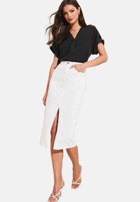 Blusa nera con scollo a V e maniche corte abbinata a una gonna bianca al ginocchio con una fessura frontale. Modella che indossa sandali trasparenti con tacco.