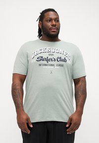 Gaiši zaļš īsām piedurknēm T-krekls, izgatavots no mīkstas auduma. Iekļauj baltu un tumši zilu tekstu: "Jack & Jones Surfer’s Club" un citus zīmola elementus.
