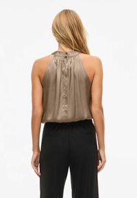 Brun sateng ermeløs topp med samlet bak, knappdetalj ved nakken, kombinert med svarte bukser. Glatt tekstur og avslappet passform.