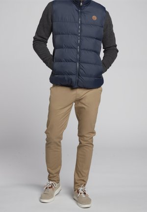 Mann trägt marineblaue gesteppten Weste über dunkelgrauem Pullover, beigefarbenen Hosen und beigen Sneakers mit weißen Schnürsenkeln, steht mit Händen in den Taschen.