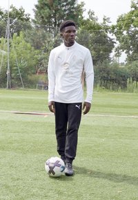 Weißes Langarmhemd mit Logo, schwarze Hose und schwarze Fußballschuhe; stehend auf Gras mit einem schwarz-weißen Fußball.