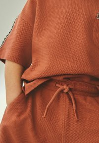 Top de tejido texturizado en color óxido, con un ajuste holgado, mangas cortas y un bolsillo lateral, combinado con pantalones a juego con cordón ajustable.