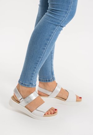 Weiße Sandalen mit silbernen Akzenten, ausgestattet mit verstellbaren Riemen und einer geschwungenen Sohle. Getragen mit blauen Skinny-Jeans, die das moderne Design hervorheben.