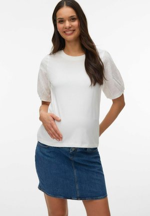 Smilende kvinde med langt brunt hår iført en hvid bluse med hulmønstrede, puffede ærmer og en blå denim nederdel, hånd hvilende på maven.