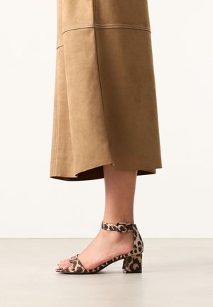 Benen met een bruine midi-rok gecombineerd met sandalen met blokhak en luipaardprint en enkelbandjes op een effen vloer.