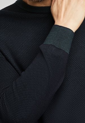Dunkles Sweatshirt mit einem strukturierten Muster, grünen gestreiften Bündchen und einem gerippten Halsausschnitt. Nahaufnahme von Stoff- und Handdetails sichtbar.