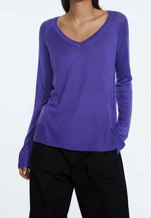 Femme portant un pull violet à manches longues avec col en V et un pantalon noir, une main dans la poche, montrée du cou jusqu'aux hanches.
