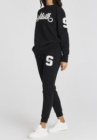 Czarna bluza z kapturem z haftem "SikSilk" w białym kolorze, zestawiona z czarnymi dresami z haftowanym "S." Białe sneakersy dopełniają stylizację.