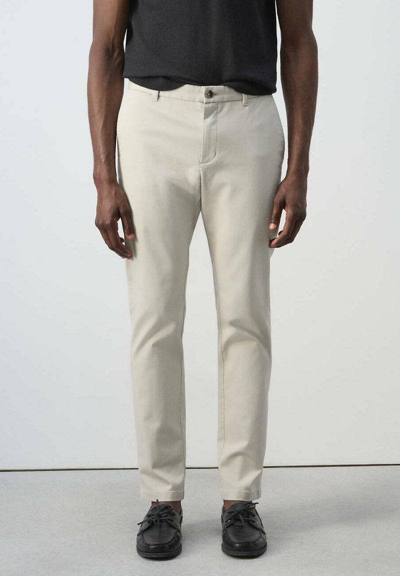 Homme portant un pantalon beige coupe droite, une chemise noire et des chaussures décontractées sombres à lacets, debout devant un fond clair uni.