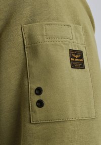 Tissu de sweat-shirt olive avec une finition texturée, comportant une poche à rabat et des boutons-pression noirs. Étiquette de la marque visible sur la poche.