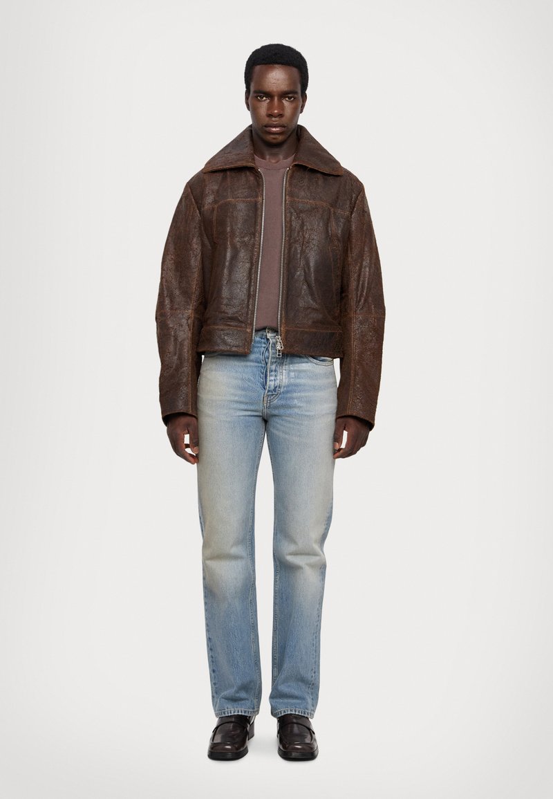 Brown leather jacket with large collar, zip closure, and textured surface, paired with light blue straight-leg jeans and dark shoes.
