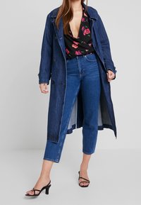 Jeans trench coat, mörkblå, med stora knappar, en lös passform, lager på lager över en blommig svart topp och högmidjade blåjeans, tillsammans med svarta klackar.