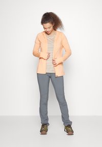 Fleece com zíper na cor laranja clara, com um acabamento texturizado, usado sobre uma camiseta listrada em cinza e branco, combinado com calças cinzas e ténis verde-escuro.