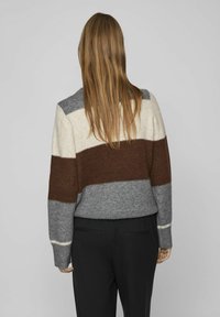 Sweter w blokach kolorystycznych w odcieniach szarości, kremu i brązu. Dzianinowa faktura, okrągły dekolt i ściągacz na końcach. Połączony z czarnymi spodniami. Widok z tyłu pokazany.