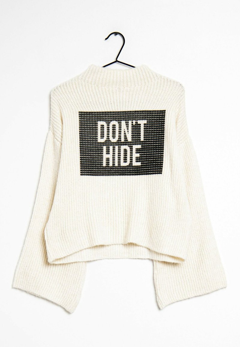 Pull blanc en tricot suspendu sur un cintre noir avec des manches larges et un patch rectangulaire noir portant l'inscription "DON'T HIDE" au dos.