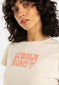 Crème-kleurig t-shirt met het "BILLABONG" logo in koraal, met palmbomenaccenten. Gemaakt van zachte, ademende katoen.