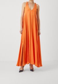 Longue robe maxi orange avec un décolleté en V profond, des bretelles à nouer réglables, un tissu fluide et des fentes sur les côtés. Portée avec des chaussures à talons hauts bleu marine.