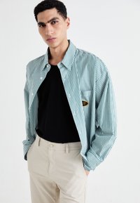 Stripet knappeskjorte i teal og hvit, med en lomme med et logo. Brukt over en svart t-skjorte og beige bukser. Glatt tekstur.