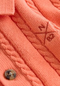 Tissu tricoté couleur corail avec des motifs torsadés, un bouton en écaille de tortue et des lettres brodées "a" et "z" croisées par une ligne diagonale.