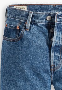 Jeans de denim azul claro con un corte recto clásico, que presentan remaches de cobre, una bragueta de botón y costuras naranjas, incluyendo un bolsillo en la parte frontal.