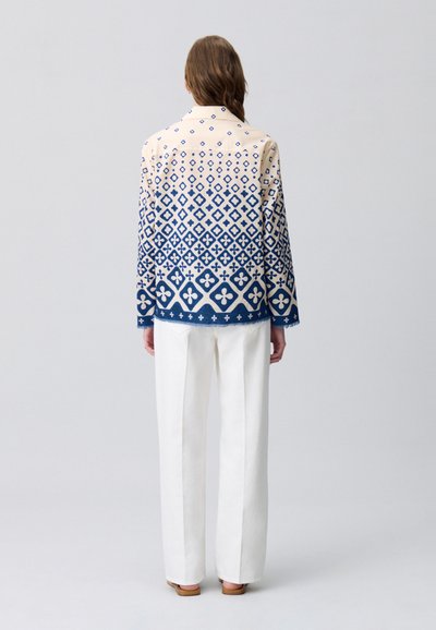 Camicia a maniche lunghe a fantasia in crema e blu navy con design geometrici. Abbinata a pantaloni a gamba larga bianchi e sandali bassi.