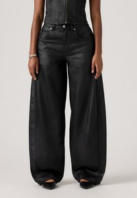 Femme portant un pantalon en cuir noir à taille haute et à jambes larges, associé à des chaussures noires à bout pointu et un haut sans manches noir avec fermeture éclair.