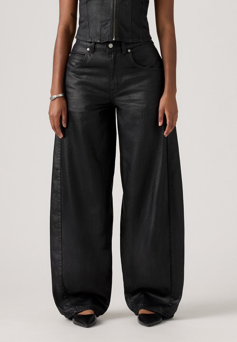 Femme portant un pantalon en cuir noir à taille haute et à jambes larges, associé à des chaussures noires à bout pointu et un haut sans manches noir avec fermeture éclair.
