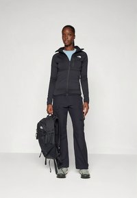 Črna trenirka z zadrgo, svetlo modra srajca, črne hlače s žepi in sive pohodniške čevlje. Črn nahrbtnik z nastavljivimi trakovi.