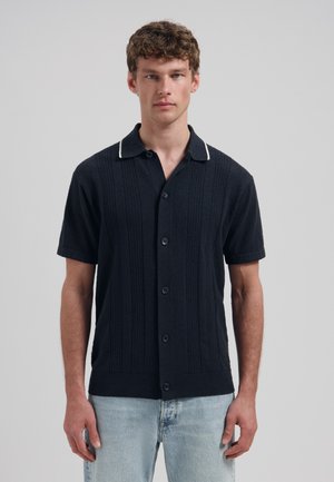 Jonge man die een zwart gebreid shirt met korte mouwen en knopen draagt, gecombineerd met lichtblauwe jeans, staand tegen een effen grijze achtergrond.