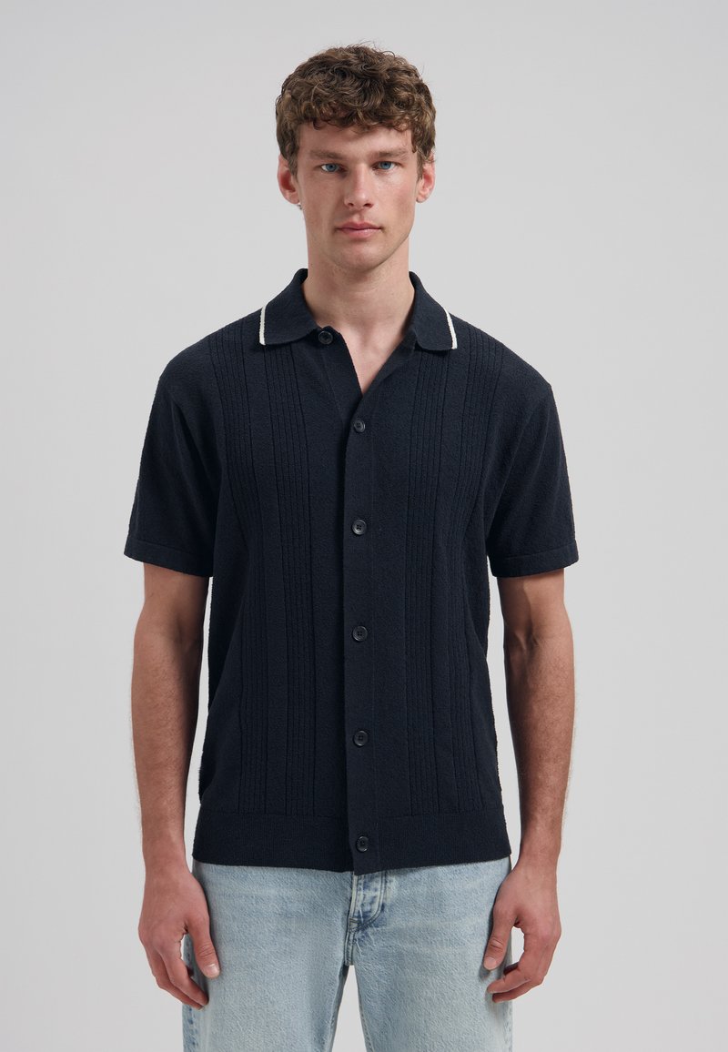 Jeune homme portant une chemise en maille noire à manches courtes avec des boutons et un jean bleu clair, debout devant un fond gris uni.
