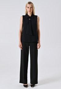 Donna bionda che indossa un top senza maniche nero con dettagli di annodatura frontale, pantaloni neri a gamba larga e scarpe nere con punta su sfondo chiaro uniforme.