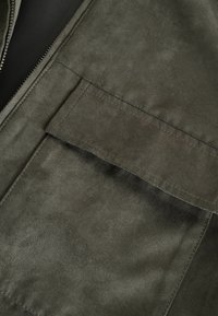 Veste zippée de couleur vert olive avec une texture douce ressemblant à du daim, dotée d'une grande poche à rabat et de détails de couture visibles.