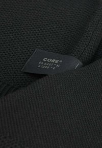 Tissu tricot noir avec une texture fine, comportant une étiquette marquée "CORE" et des coordonnées géographiques. Pris sous un angle pour montrer les plis.