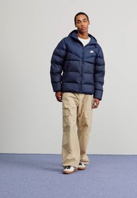 Casaco puffer azul-marinho com capucho, apresentando um fecho com zíper e painéis cosidos. Usado sobre calças cargo bege e ténis brancos.