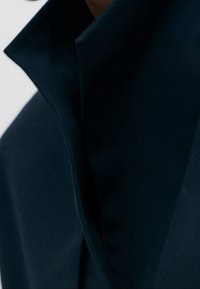 Ciemnoniebieska kurtka z wysokim, usztywnionym kołnierzem. Materiał wygląda na gładki z delikatnym połyskiem. Wyraźne detale przeszycia wzdłuż krawędzi.