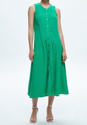 Zielona sukienka midi bez rękawów zapinana na guziki z szwem w kształcie litery V, noszona z białymi sandałami na obcasie z paskiem wokół kostki.